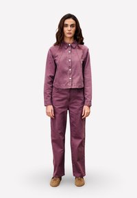 Femme debout, portant une veste en denim violette assortie avec des boutons blancs et un pantalon taille haute, associée à des chaussures beiges, sur un fond neutre.