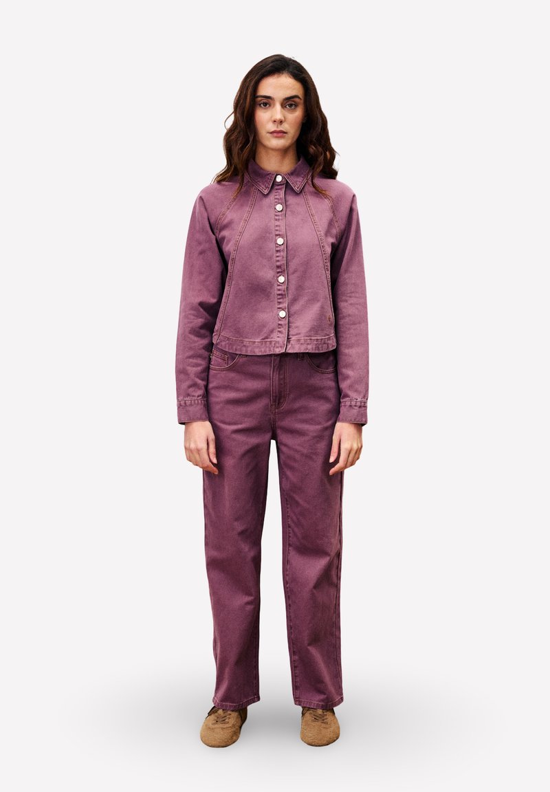 Femme debout, portant une veste en denim violette assortie avec des boutons blancs et un pantalon taille haute, associée à des chaussures beiges, sur un fond neutre.