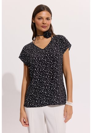 Kobieta ubrana w czarną bluzkę z krótkim rękawem i dekoltem w serek w białe groszki, białe spodnie, czarną apaszkę w groszki na szyi oraz białą bransoletkę z koralików.