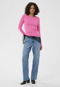 Mujer con una blusa de manga larga rosa brillante, jeans azules con detalles de botones y botas de tobillo negras con tacón, de pie contra un fondo claro y liso.