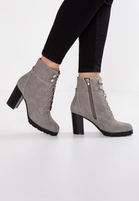 Bottes cheville en daim gris avec talons carrés, fermeture éclair latérale et devant à lacets, portées avec un pantalon noir ajusté sur fond blanc.