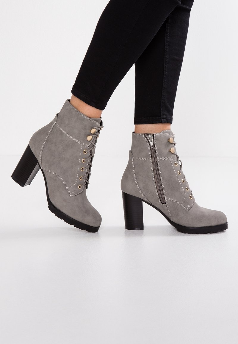 Bottes cheville en daim gris avec talons carrés, fermeture éclair latérale et devant à lacets, portées avec un pantalon noir ajusté sur fond blanc.