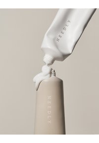 Vit tub med etiketten "NEEDLY" som pressar ut en krämig substans på en slät taupe-färgad flaska, båda med minimalistisk design.