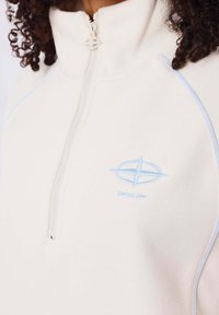 Felpa leggera color crema con zip a quarto e cuciture di accento blu, logo ricamato. Texture morbida con colletto alto e zip standard.