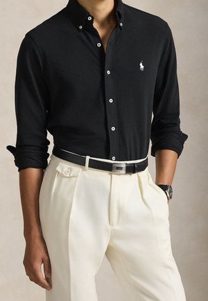Homme portant une chemise noire à manches longues boutonnée avec un logo blanc, un pantalon blanc plissé, une ceinture noire et une montre-bracelet, mains détendues sur les côtés.