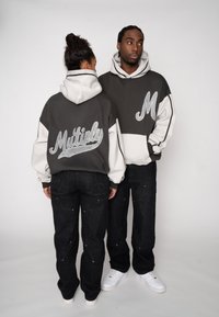 Hanorace gri și albe cu broderie texturată „Multiply” și „M”. Amândoi poartă blugi închiși la culoare cu cusături decorative și adidași albi.