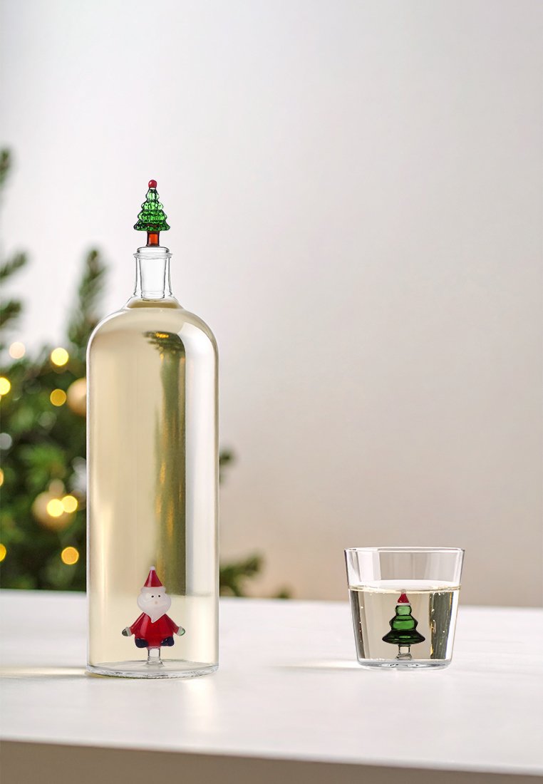 Przezroczysta szklana butelka z zielonym korkiem w kształcie choinki, w środku której znajduje się figurka Mikołaja w czerwonym ubraniu. Obok stoi szklanka z zielonym wzorem choinki.