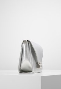 Stříbrná metalická kožená kabelka ve tvaru psaníčka s klopou a kovovým zapínáním, vystavená na bílém podstavci proti jednoduchému světlému pozadí.