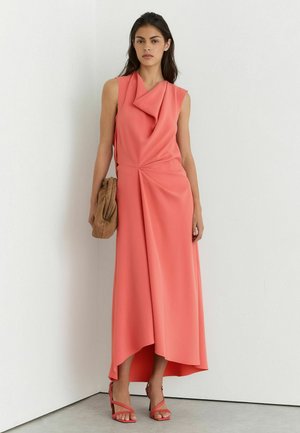 Frau in korallenfarbenem ärmellosem Kleid mit asymmetrischem Saum, hält eine beige Clutch, trägt korallenfarbene Riemchenabsätze, vor einer weißen Eckwand.