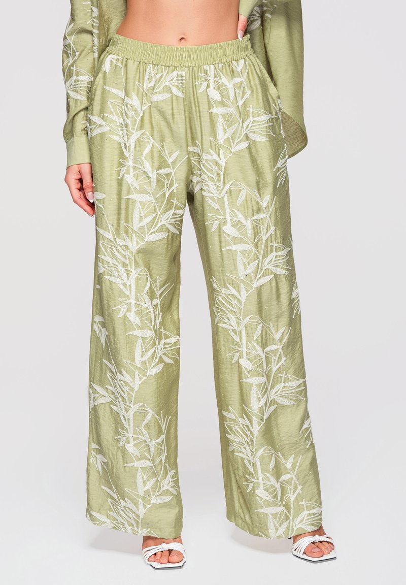 Pantalon large vert clair à imprimé botanique blanc, taille élastique, porté avec un haut assorti et des sandales blanches à bout ouvert.