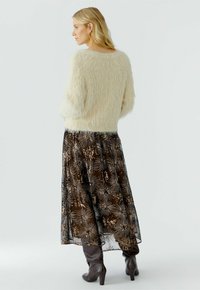 Eine Frau mit blonden Haaren trägt einen cremefarbigen, flauschigen Pullover, einen langen schwarzen Rock mit metallischen Sonnenblumenmustern und dunkelbraune Stiefeletten mit Absatz, während sie steht.