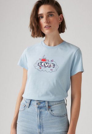 Kvinna med kort brunt hår bär en ljusblå Levi's T-shirt med en grafik av körsbär och blommor, kombinerat med högmidjade ljus denimjeans.