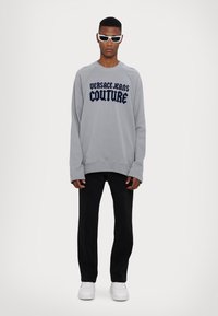 Šedá mikina s námorníckym modrým textom "VERSACE JEANS COUTURE", spárovaná s čiernymi nohavicami a bielymi teniskami. Model má na sebe biele slnečné okuliare.