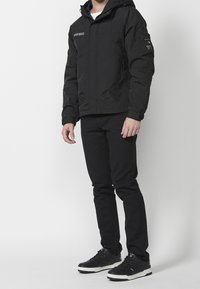 Svart isolerad jacka med huva, dragkedja framtill, sidofickor och broderad logga. Buren med svarta tajta byxor och svarta sneakers.