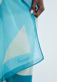 Bufanda translúcida en tonos de azul, con un diseño geométrico. Los bordes están cosidos, con el logo "Triumph" en elegante tipografía en la parte inferior.
