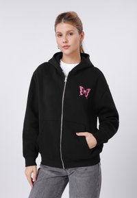 Sweat à capuche noir zippé en coton, orné d'une broderie de papillon rose sur la poitrine gauche. Capuche ajustable et poches avant.