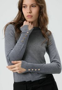 Grijs topje met lange mouwen, een geribbelde halslijn en gouden knoopaccenten op de manchetten, gemaakt van een zacht, gestructureerd gebreid materiaal.