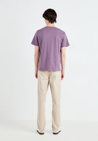 T-shirt di cotone viola con maniche corte e collo tondo. Abbinato a pantaloni beige chiaro e sneakers bianche, mostrando un fit rilassato.