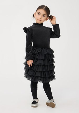 Abito nero con maniche lunghe texturizzate, corpetto arricciato e gonna in tulle a strati. Calze in maglia nere e scarpe argento con dettaglio a fiocco.