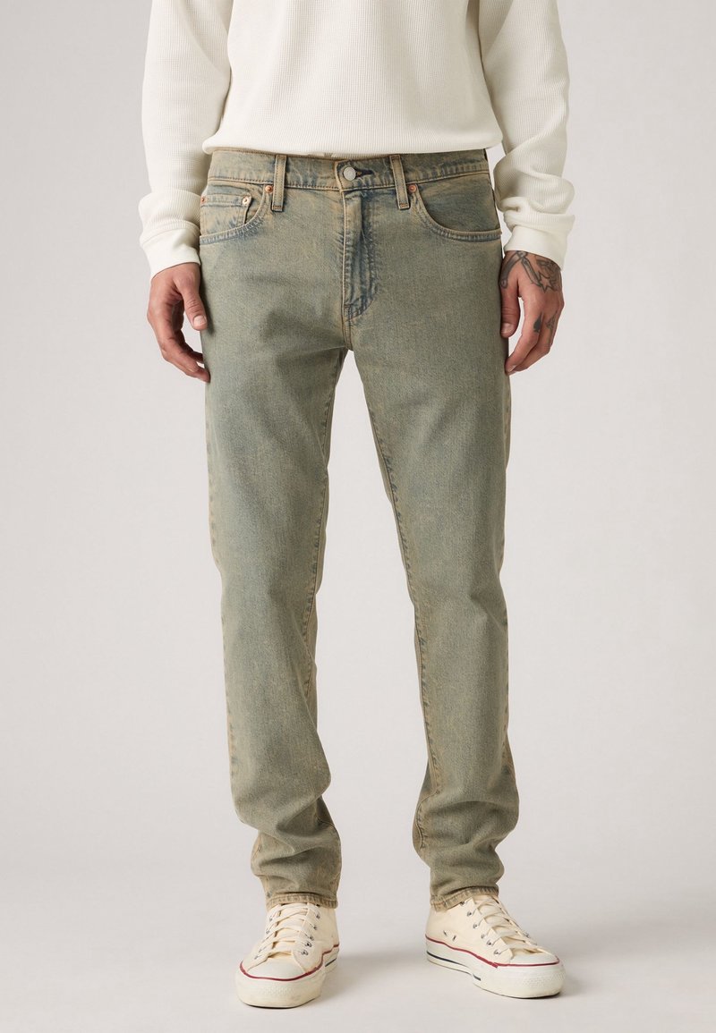 Personne portant un jean marron clair, des baskets blanches et une chemise blanche à manches longues, debout les bras détendus le long du corps.