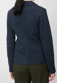 Chaqueta texturizada azul marino con un diseño ajustado y cuello entallado. Cuenta con mangas largas y una tela suave, combinada con pantalones verde oliva.