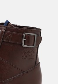 Botín de cuero marrón con interior azul, detalle de hebilla plateada y logotipo de marca grabado, con acentos de costura texturizada.
