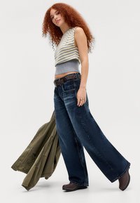 Woman with curly red hair wearing striped sleeveless top, wide-leg jeans, brown belt, and boots, holding an olive green jacket while walking.