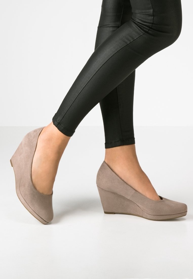 Jambes portant des leggings effet cuir noir avec des talons compensés en daim beige, un pied légèrement levé sur un fond de sol blanc.