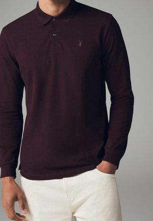 Homme portant un polo à manches longues de couleur bordeaux foncé avec un petit logo brodé sur la poitrine et un pantalon blanc, une main dans la poche.