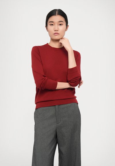 Red knitted sweater with long sleeves and a round neckline, paired with gray patterned trousers. Smooth texture, classic fit.