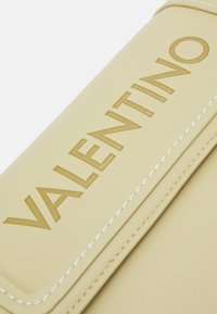 Beige läderhandväska med präglad "VALENTINO"-logotyp, vit sömnad och slät yta. Har en strukturerad design och rundade kanter.