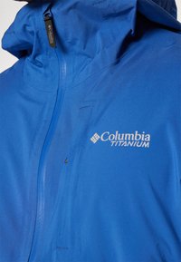 Niebieska kurtka Columbia Titanium z zamkiem błyskawicznym z przodu, wysokim kołnierzem i logo na piersi. Gładka tekstura i lekki materiał.