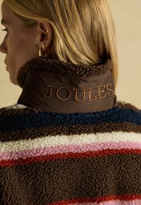 Braune Fleecejacke mit mehrfarbigen Streifen in Creme, Pink, Rot, Braun und Marineblau. 'JOULES' ist auffällig auf dem Kragen gestickt.