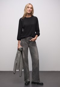 Czarna bluzka z długim rękawem zestawiona z jasnoszarymi, szerokimi spodniami z wysokim stanem. Trzyma szarą kurtkę, nosi czarne buty. Gładka faktura tkaniny.