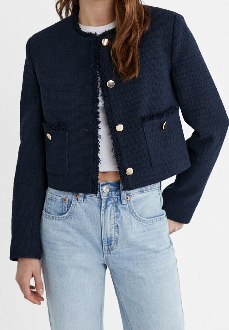 Veste courte texturée bleu marine avec bords effilochés, boutons dorés et deux poches avant. Associée à un jean taille haute bleu clair.