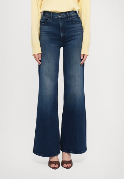 Vaqueros acampanados de denim azul oscuro con una cintura alta. Presenta cinco bolsillos y una textura suave, combinados con tacones abiertos marrones.