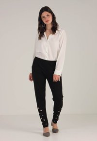 Fehér, hosszú ujjú blúz gomb részletekkel, fekete nadrággal párosítva, amelynek szegélyén kör alakú díszek találhatók. Szürke balerina cipők teszik teljessé az összeállítást.