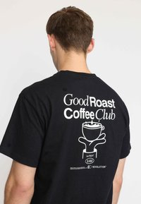Zwarte katoenen T-shirt met witte grafische tekst: "Good Roast Coffee Club" en een illustratie van een koffiekop, met een klein label onderaan.
