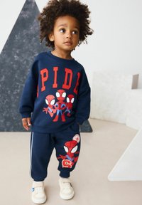 Toddler with curly hair wearing navy blue Spider-Man sweatshirt and pants, standing indoors near geometric shapes.