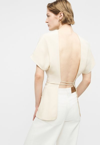Femme aux cheveux blonds courts portant un haut crème à dos ouvert avec des manches courtes, noué avec des fines bretelles, associé à un pantalon blanc taille haute.
