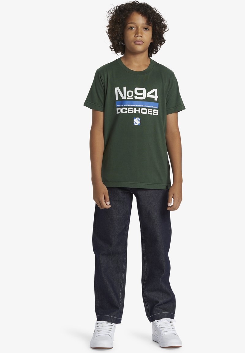 Zelené bavlněné tričko s bílým a modrým potiskem "No 94 DC Shoes". V kombinaci s tmavě modrými džíny a bílými teniskami.