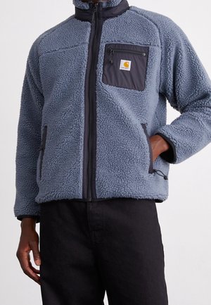 Mężczyzna w szarej polarowej kurtce z czarnym zamkiem błyskawicznym, czarną kieszenią na piersi z logo i rękami w bocznych kieszeniach, na jednolitym tle.