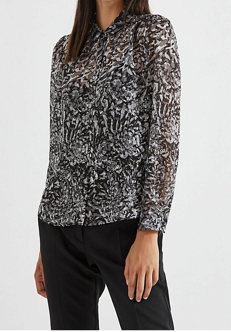 Blusa nera trasparente con motivo floreale in tonalità di grigio, con chiusura a bottoni sul davanti, colletto e maniche lunghe, abbinata a pantaloni neri.