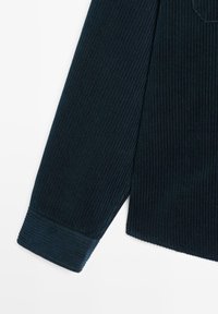 Manica di camicia in corduroy blu scuro con texture a coste verticali, dotata di dettagli cuciti su polsini e orlo. Finitura liscia sul tessuto.