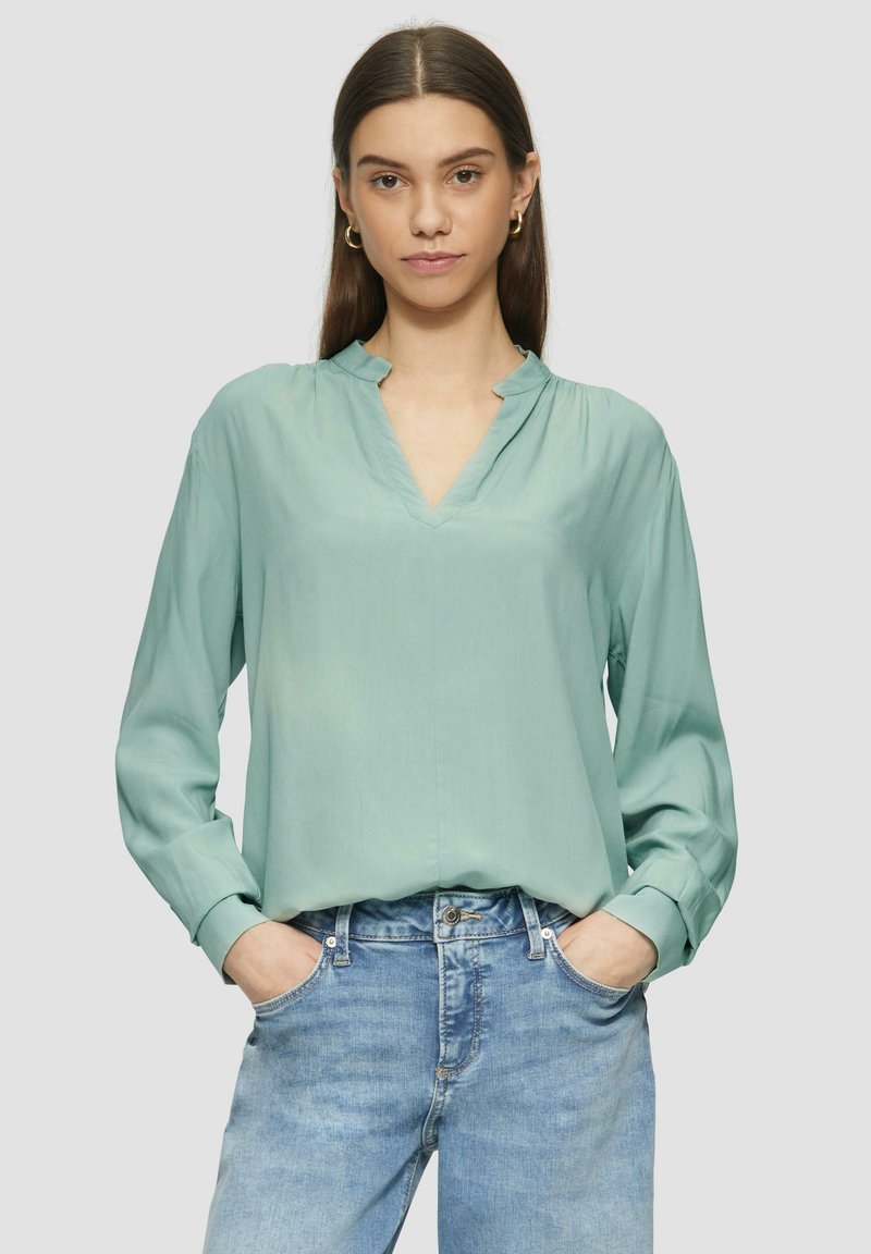 Mintgrünes Blusenshirt mit V-Ausschnitt und leichter Kragen, langen Ärmeln und weicher Textur, kombiniert mit hellblauen Jeans.
