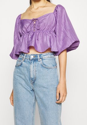 Woman wearing a purple, puff-sleeve crop top with front lace-up detail and light blue high-waisted jeans.