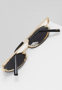 Gafas de sol con marco de metal dorado y lentes circulares negros, con patas de acetato negro y almohadillas nasales transparentes ajustables.