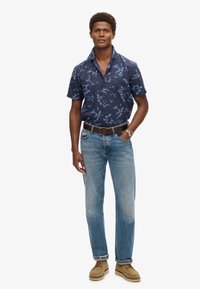 Camisa de manga corta de color navy con un patrón floral azul, con cuello abotonado, combinada con jeans de pernera recta de color azul claro y zapatos de ante color tostado.