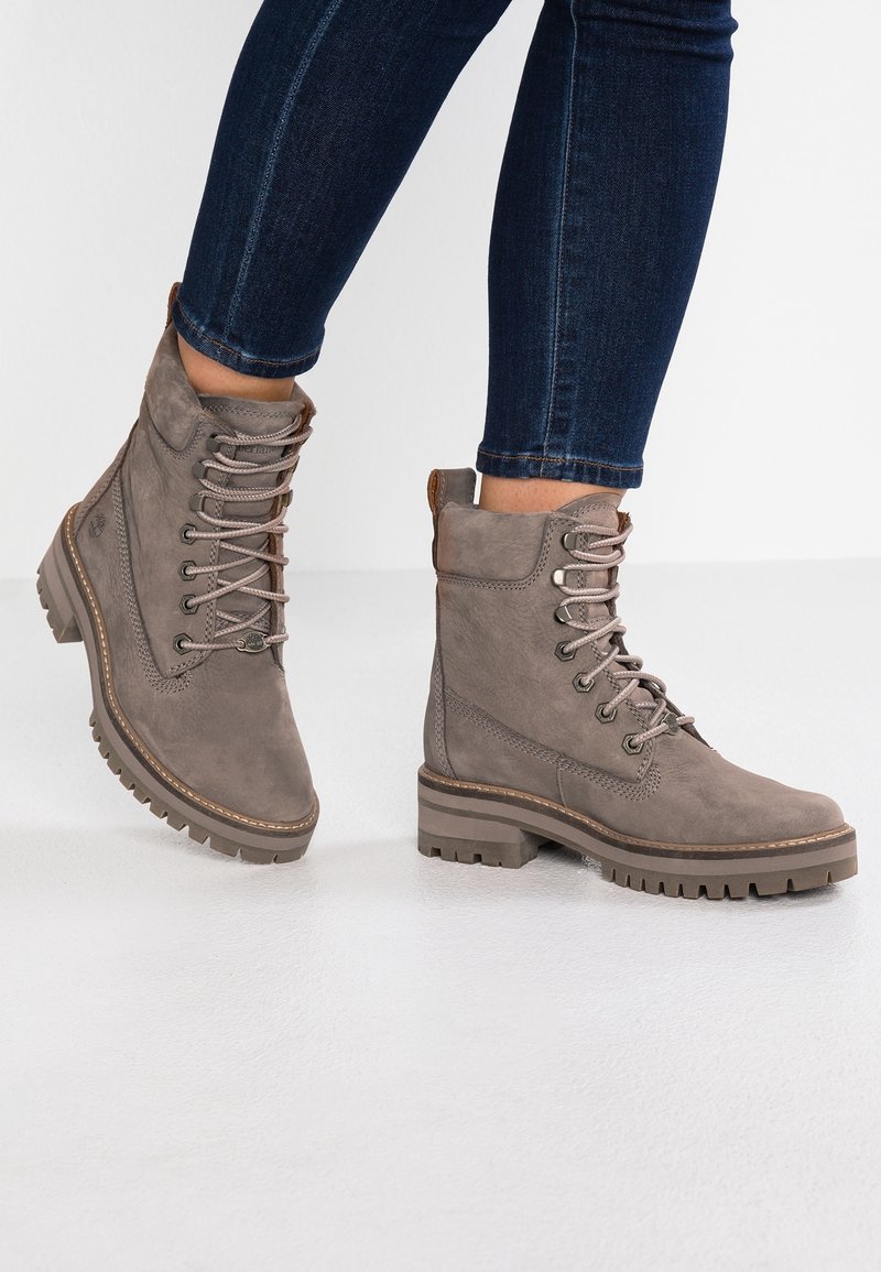 Harmaat nahkasaappaat nilkkamittaiset, joissa on nauhat, teksturoitu pinta ja kestävä kumipohja. Varustettu kohotetulla kantapäällä ja oranssilla yksityiskohdalla takana.