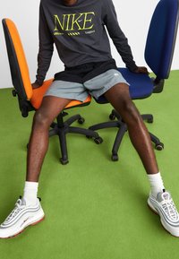 Schwarzes Langarm-Nike-Shirt mit gelbem Logo, graue Shorts, weiße Nike Air Max 97 Schuhe, sitzend auf orangefarbenen und blauen Bürostühlen. Grüner Boden.